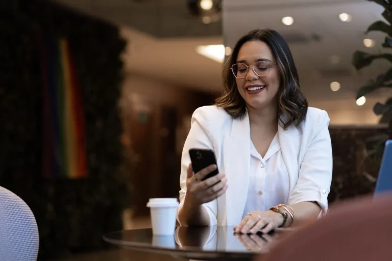 Mujer sonriente usando su smartphone en una cafetería, representando la optimización de la experiencia móvil en la gestión de viajes corporativos.