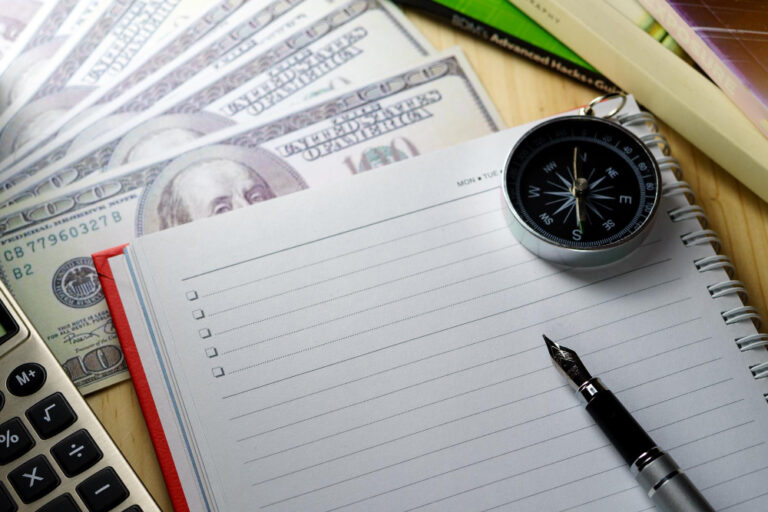 Cuaderno con lista de verificación (checklist), pluma fuente, brújula y billetes de dólar, representando la creación de políticas de gastos de viaje y control de viáticos empresariales.